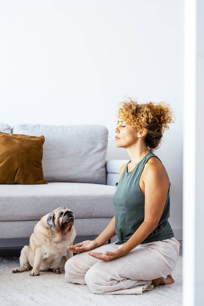  healthy adult woman at home doing meditation exercise in yoga oriental zen position with her best friend dog pug sitting near her looking curious. Indoor leisure activity people in apartment - Photo, Image
