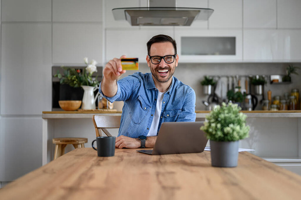 Портрет щасливого бізнесмена, що показує кредитну картку під час покупки в Інтернеті за ноутбуком за столом - Фото, зображення