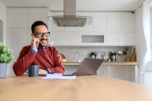Glücklicher junger Geschäftsmann, der mit dem Smartphone telefoniert, während er mit dem Laptop auf dem Schreibtisch im Home Office sitzt - Foto, Bild