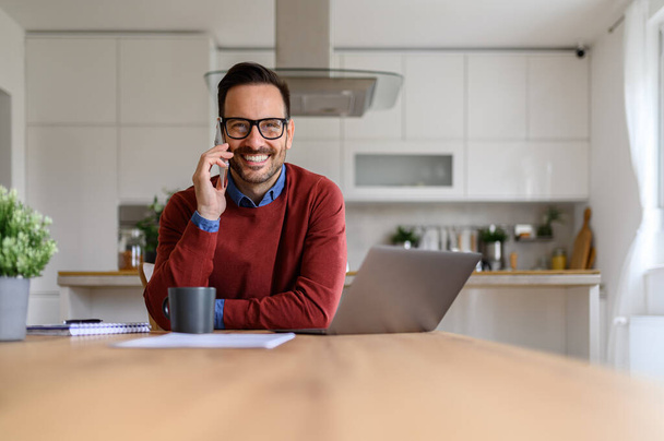 Männlicher Unternehmer telefoniert mit dem Handy und schaut in die Kamera, während er zu Hause über Laptop arbeitet - Foto, Bild
