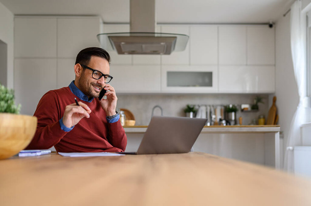 Freelancer bonito segurando caneta e discutindo por telefone celular enquanto trabalhava na mesa no escritório em casa - Foto, Imagem