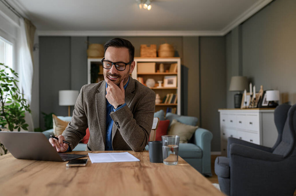 Elegantní mladý podnikatel s rukou na bradě myšlení při práci nad notebookem na stole doma - Fotografie, Obrázek