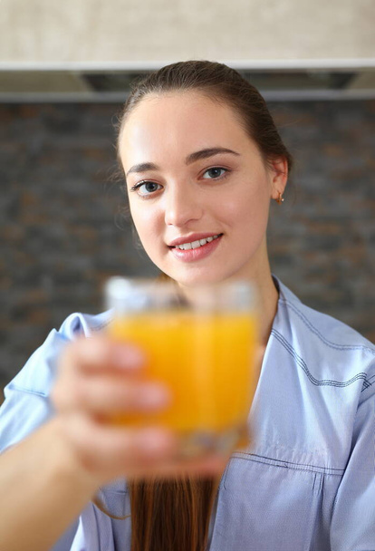 Молодая красивая привлекательная девушка в пижаме на кухне пьет апельсиновый сок держа стакан в руке
 - Фото, изображение