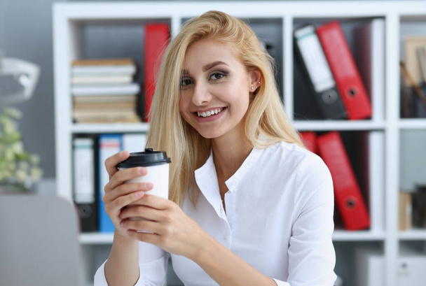 Lächelnde Geschäftsfrau, die aus einer Papptasse im Büro Kaffee trinkt und in die Kamera blickt. In der Hand halten und während einer Pause ausruhen. - Foto, Bild