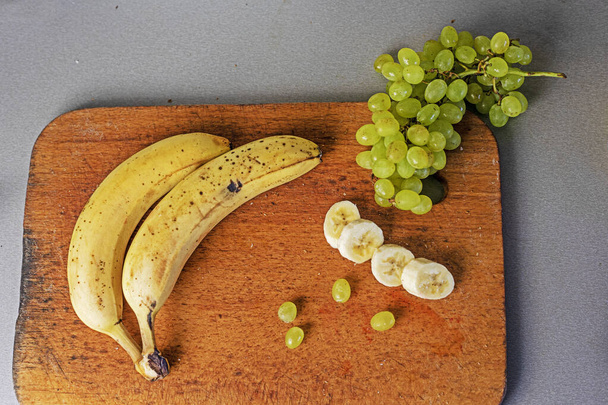 Banda bananów z bandą zielonych winogron na drewnianej desce do krojenia na szarym tle. płaskowyż - Zdjęcie, obraz