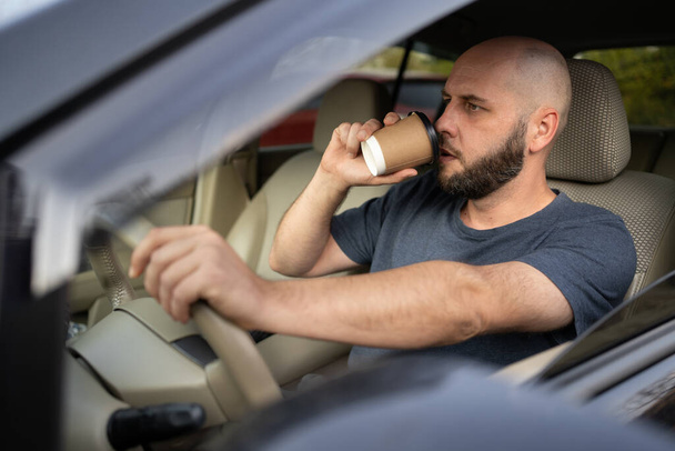 Mann fährt Auto, während er Kaffee trinkt. Er blickt in die Kamera. Transport-, Fahrzeug- und Personenkonzept - Foto, Bild