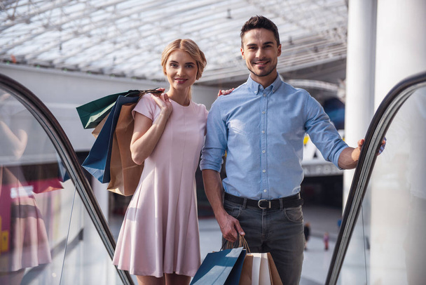 schönes Paar mit Einkaufstaschen blickt in die Kamera und lächelt, während es auf der Rolltreppe im Einkaufszentrum steht - Foto, Bild