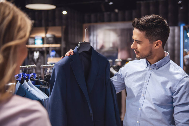Casal bonito está escolhendo terno formal e sorrindo ao fazer compras na loja masculina
 - Foto, Imagem