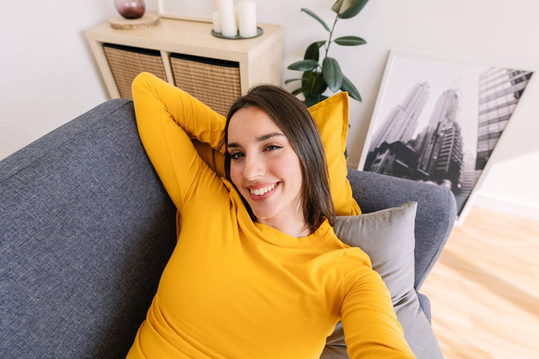 幸せな若い美しい女性は自宅でソファーに横たわっているカメラで微笑んでいる間,自撮りの肖像画を撮ります - 写真・画像