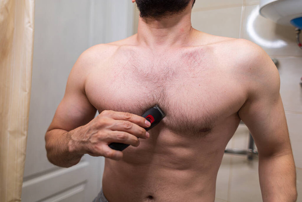 Un hombre quita el pelo no deseado de su cuerpo usando una afeitadora eléctrica. Un hombre con una figura atlética en el baño. - Foto, imagen