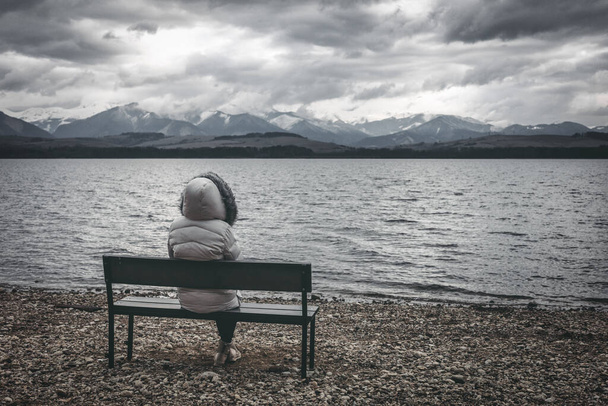 Vrouw in jasje zittend op een bankje aan de oever van het meer. Donkere bewolkte lucht met bergen op de achtergrond - Foto, afbeelding