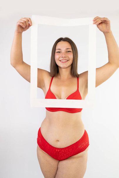 Body positivity and self love concept. Waist up shot of joyful body positive young woman holding frame feels confident love own body wears red lingerie poses over white wall - Photo, Image
