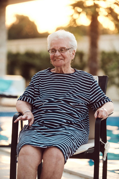 Niefiltrowany portret, prawdziwa starsza kobieta z wdziękiem siedzi na krześle, pokazując autentyczność starzenia się ze zmarszczkami i naturalną twarzą.  - Zdjęcie, obraz
