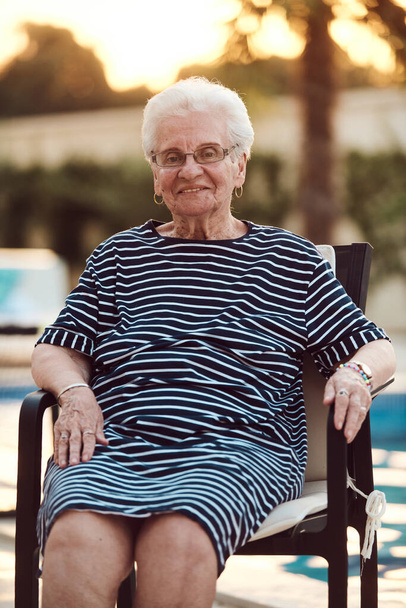 Niefiltrowany portret, prawdziwa starsza kobieta z wdziękiem siedzi na krześle, pokazując autentyczność starzenia się ze zmarszczkami i naturalną twarzą.  - Zdjęcie, obraz
