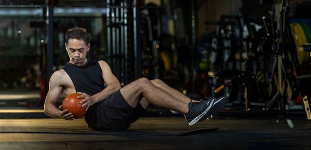 Asijské muž je cvičení v tělocvičně dělá sedící šikmé twist cvičení s váhou medicíny míč na podlaze, které posilují břišní a svalové hmoty   - Fotografie, Obrázek