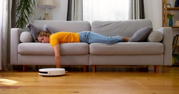 Hyvin väsynyt nainen nukkuu sohvalla kotona, kun robotti pölynimuri työskentelee lattialla. Laadukas 4k kuvamateriaalia - Materiaali, video