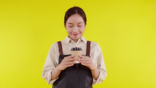 Portrait of young asian barista woman wearing apron smell coffee cup on yellow background, waitress or entrepreneur holding coffee with excited, small business or startup, waiter of cafe. - Footage, Video