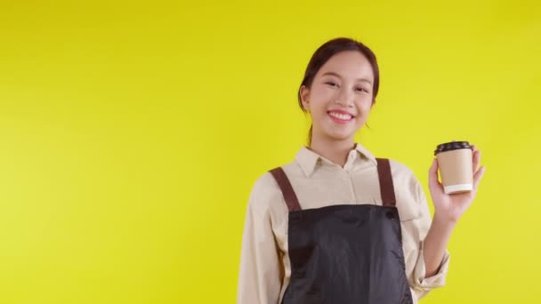 Portré fiatal ázsiai barista nő visel kötény gazdaság kávéscsésze és bemutató sárga háttér, pincérnő vidám és bemutató és hüvelykujj fel, kisvállalkozás vagy startup, pincér kávézó. - Felvétel, videó