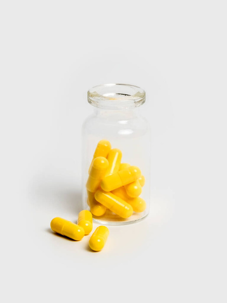 Keltainen pillereitä lähellä pysyvän avattu lasipullo eristetty valkoisella pohjalla. Käsitys lääkehoidosta, vitamiineista tai lisäravinteista kapseleissa, terveydenhuollossa tai huumeriippuvuudessa. - Valokuva, kuva