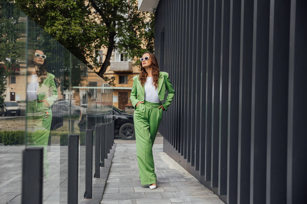 Mujer confiada elegante de moda que usa gafas de sol de moda rosa, pañuelo de seda, chaqueta de traje, con bolsa verde, posando en la calle de la ciudad europea. Retrato de moda al aire libre. Copiar, espacio vacío para texto - Foto, imagen