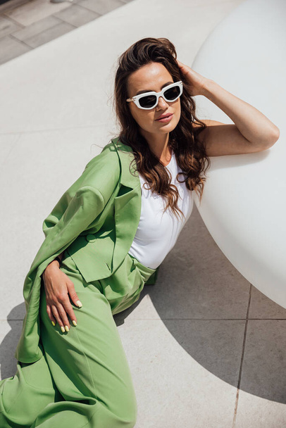 Mujer confiada elegante de moda que usa gafas de sol de moda rosa, pañuelo de seda, chaqueta de traje, con bolsa verde, posando en la calle de la ciudad europea. Retrato de moda al aire libre. Copiar, espacio vacío para texto - Foto, Imagen
