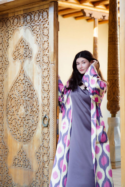 Beautiful fashion model, girl in traditional Uzbekistan fashion dress with ikat pattern open door. Tashkent, Uzbekistan - Photo, Image