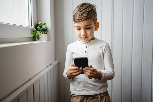 one boy caucasian child preschooler hold smartphone mobile phone at home play video games childhood and growing up technology addiction concept use smartphone app for online browsing or watch video - Foto, afbeelding