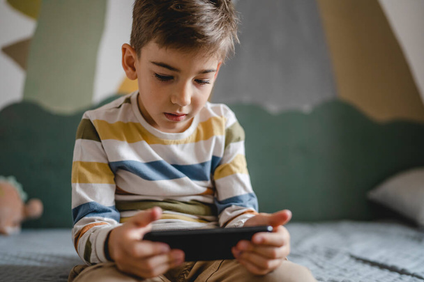 one boy caucasian child preschooler hold smartphone mobile phone at home play video games childhood and growing up technology addiction concept use smartphone app for online browsing or watch video - Fotoğraf, Görsel