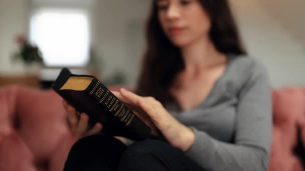 Een langharig meisje die alleen thuis een boek leest, zittend op de bank. Een jonge vrouw leest de Bijbel. Een vrouw houdt een bijbel in haar handen en bestudeert het woord van God. - Video