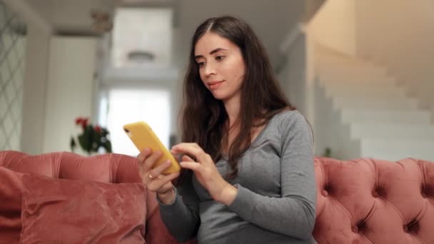 Χαρούμενη νεαρή γυναίκα που χρησιμοποιεί smartphone ενώ κάθεται στον καναπέ. Ελκυστική χαμογελαστή γυναίκα που χρησιμοποιεί την εφαρμογή στο κινητό τηλέφωνο στο σπίτι. Όμορφη κοπέλα χαλαρώνει ενώ συνομιλεί στο κινητό τηλέφωνο στο σπίτι - Πλάνα, βίντεο
