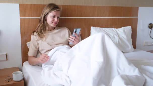 Young woman using smartphone in hotel bed after waking up - Footage, Video