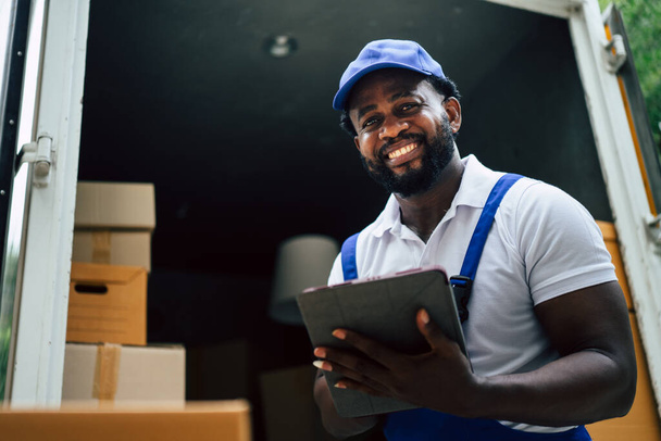 African courier man house mover Comprobación de caja de cartón con tableta antes de pasar al cliente. Hombre optimista auténtico feliz y disfruta de su trabajo. - Foto, imagen