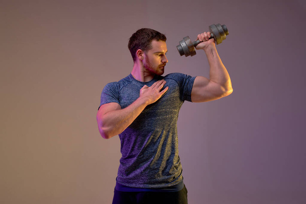serious fit man performing weightlifting exercise. guy looks at his arm . isolated brown background. - Foto, Imagem