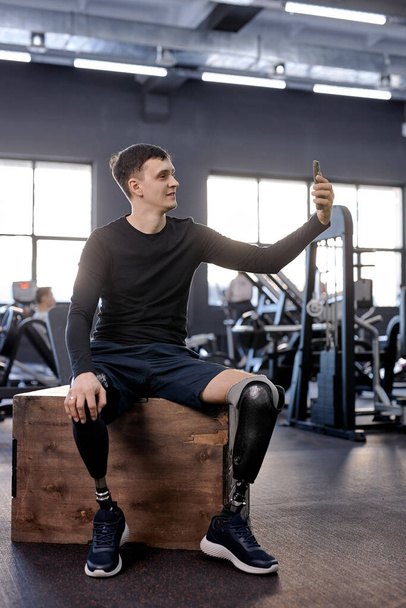 sportsman making video phone call. guy sitting on wooden box, taking selfie at gym, full length photo. disabled man taking photo at sport center. love of life. blogger shooting video - Fotografie, Obrázek