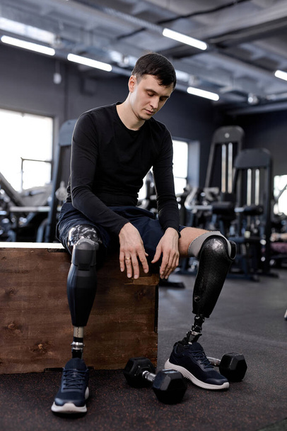 sportsman is tired , sitting on the box, looking at dumbbells on the floor. full length photo .tiredness, modern sport equipment in the background of the photo - Zdjęcie, obraz