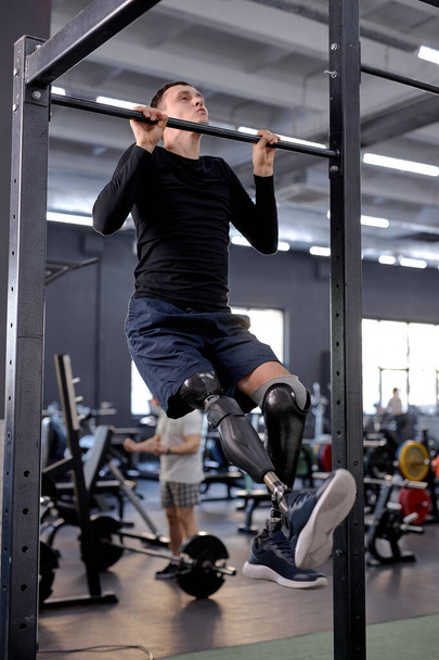 active ambitious man with prosthetic leg wearing sports clothing doing pull-ups on bar in modern gym, full length side view shot, free time, spare time, lifestyle - Фото, изображение