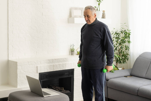 Sport in mature age. Happy middle-aged man doing stretching exercises in front of laptop at home, watching online tutorials, in living room interior, free space. - Foto, imagen