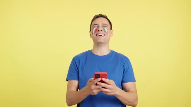 Studio video s barevným pozadím italského muže s vlajkou Španělska namalovanou na tváři, dívá se na svůj mobilní telefon s úsměvem a ukazuje ho kameře s obrazovkou v chroma klávese. - Záběry, video