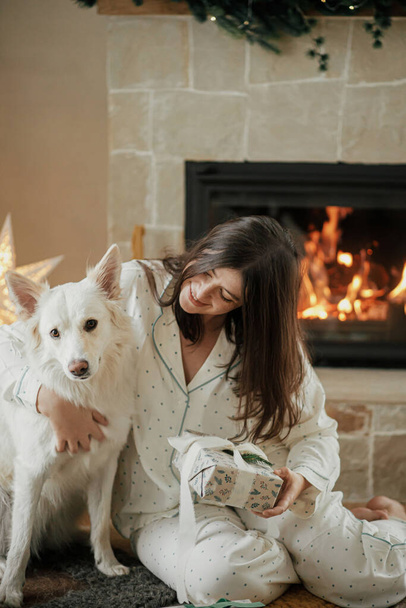 Hermosa mujer en pijama con un elegante regalo de Navidad y abrazando al lindo perro blanco en el árbol decorado y la chimenea, disfrutando de una acogedora mañana de Navidad en la sala de estar festiva. Feliz Navidad. - Foto, Imagen