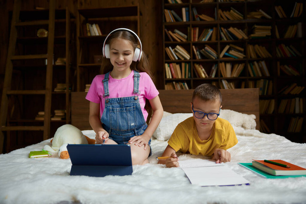 Glückliches Schulkind, das zu Hause gemeinsam lernt und Hausaufgaben vorbereitet. Kleine Schwester hört Lehrerin mit Headset und digitalem Tablet-Gerät zu, während ihr Bruder Aufgabe in Copybook schreibt - Foto, Bild