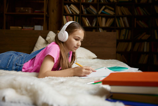 ワイヤレスヘッドフォンで自宅で勉強しているかわいい少女. 遠隔学習を楽しむウェブレッスンを持つ愛らしい子供の学生 - 写真・画像
