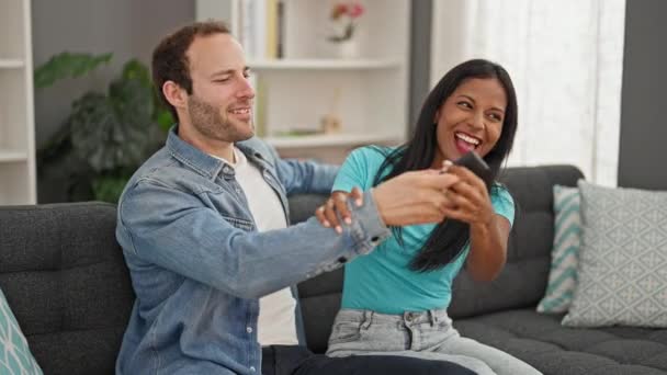 Hermosa pareja sentada en el sofá luchando por el control remoto de la televisión en casa - Metraje, vídeo