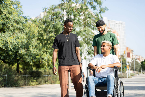 Jóvenes amigos empujando silla de ruedas del hombre discapacitado, pasándola bien juntos caminando por el parque de la ciudad - Foto, imagen
