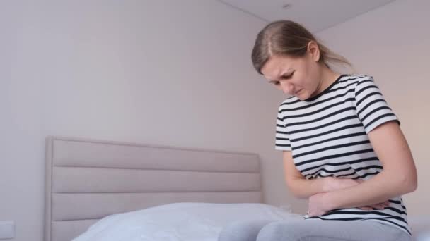 Una giovane donna malata si siede sul letto a casa, si tocca lo stomaco, soffre di dolori addominali. Ragazza si sente male, lotta con il dolore mestruale.  - Filmati, video