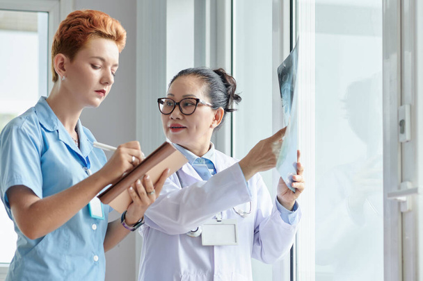 Asian professional doctor pointing at x-ray image while nurse making notes in notebook during work at office - Photo, Image