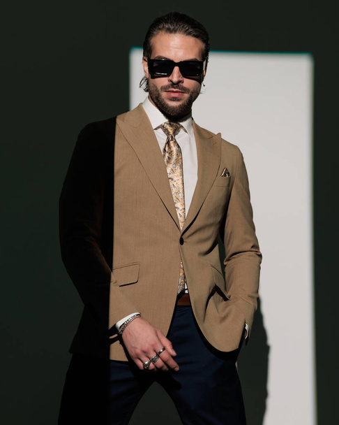 σέξι νεαρός επιχειρηματίας με γυαλιά ηλίου φορώντας υψηλής ποιότητας καφέ κοστούμι, κρατώντας το χέρι στην τσέπη και ποζάροντας σε γκρι φόντο - Φωτογραφία, εικόνα