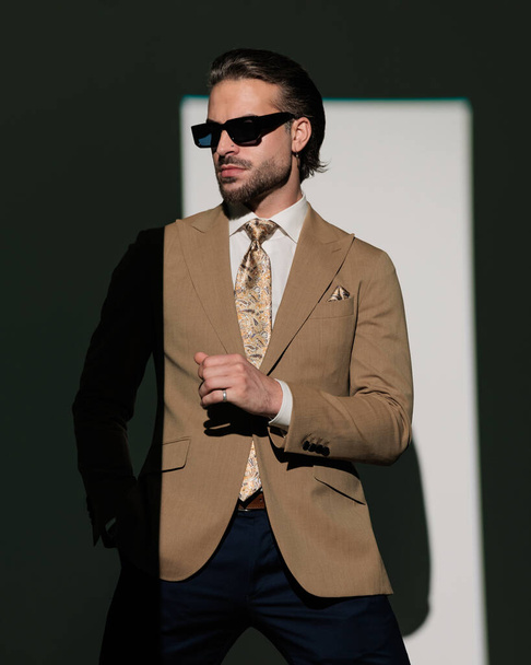 elegante zakenman met een zonnebril op zoek naar opzij en hand in hand terwijl poseren met de arm in een koele manier op grijze achtergrond - Foto, afbeelding
