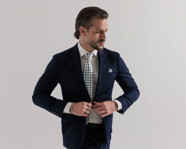 Porträt eines eleganten Geschäftsmannes der gehobenen Klasse, der einen marineblauen Anzug knöpft und vor grauem Hintergrund zur Seite schaut - Foto, Bild
