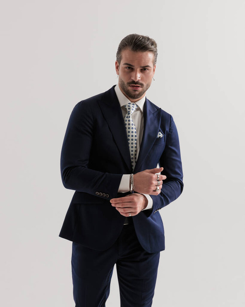 sexy businessman in navy blue suit adjusitng sleeves, looking forward and posing in front of grey background  - Фото, изображение