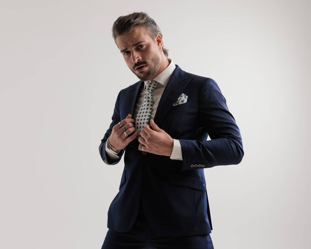arrogant elegant fashion man looking forward while fixing luxury navy blue suit in front of grey background  - Photo, Image